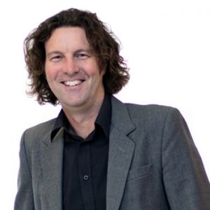 Glenn Howe in a black shift and gret suit jacket smiling at the camera.